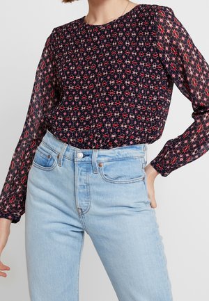 Vrouw draagt een doorschijnende lange mouwen bloemenblouse met geplooide voorkant, ingestopt in een lichtblauwe spijkerbroek met hoge taille, met hand op heup.