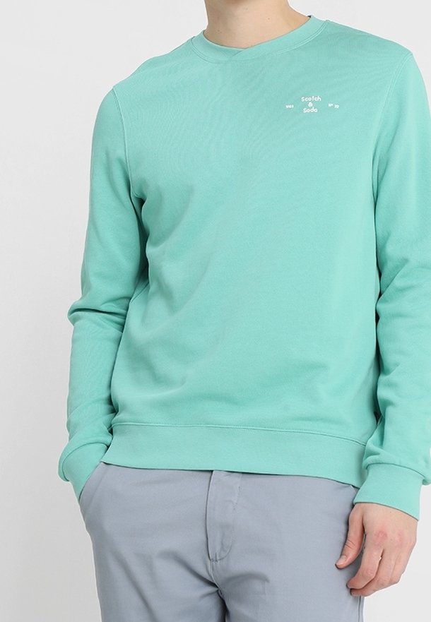 Personne portant un sweat-shirt à manches longues vert menthe avec le logo "Scotch & Soda" sur la poitrine et un pantalon gris clair, les mains partiellement dans les poches.