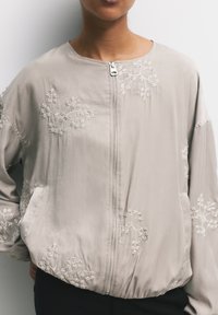 Giacca bomber grigia chiara con delicata ricamo floreale bianco, chiusura con zip e vestibilità ampia. Tessuto morbido e setoso con orlo elasticizzato.