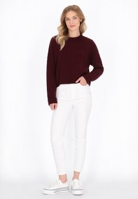 Burgundy-Strickpullover mit Rundhalsausschnitt, kombiniert mit weißen Skinny-Jeans und weißen Turnschuhen mit schwarzen Streifen auf der Sohle.