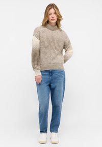 Suéter beige de punto con patrón texturizado, cuello alto y mangas en crema contrastantes. Combinado con jeans azul claro y zapatillas blancas.