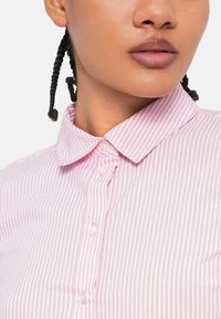 Camisa de botão com riscas rosa e brancas, com colarinho clássico. Apresenta botões e uma textura de tecido suave. Vista aproximada da gola.