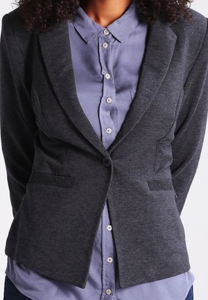 Grijze gestructureerde blazer met notch-revers, twee voorzakken en een enkele knoopsluiting, gedragen over een lichtblauw overhemd met knopen.
