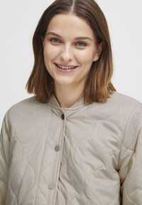 Beige vadderad jacka med rund halsringning, som har en slät yta, synliga sömmar och grå tryckknappar.