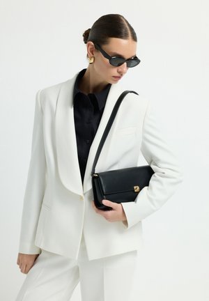 Mujer con traje blanco y camisa negra sosteniendo un bolso negro sobre el hombro, usando gafas de sol negras y pendientes de oro.