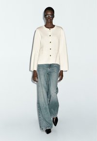 Cremefarbener Cardigan mit goldenen Knöpfen, kombiniert mit hellblauen Jeans mit weitem Bein und spitzen schwarzen Schuhen. Das Model trägt eine schwarze Brille.