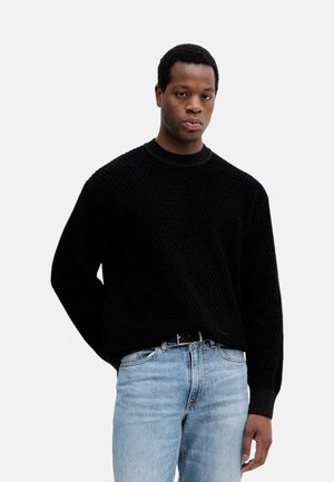 Giovane uomo che indossa un maglione nero lavorato a maglia e jeans azzurri chiari, in piedi con una mano dietro la schiena contro uno sfondo chiaro e uniforme.