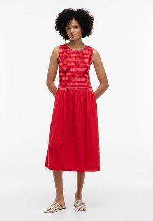 Kvinde med krøllet hår iført en ærmeløs rød midi-kjole med vandrette syninger på overdelen, kombineret med beige vævede slip-on sko.