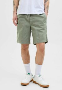Bermudas de algodón verde claro con frente plano, bolsillos laterales y un corte relajado, combinadas con calcetines blancos y zapatillas blancas con suelas marrones.