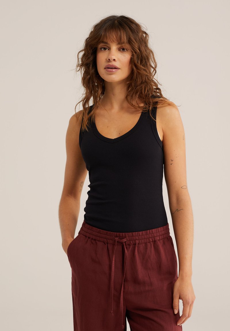 Hajlított barna hajú nő fekete ujjatlan felsőt és bordó zsinóros nadrágot viselve, zsebében tartja a kezét, és egyszerű háttér előtt áll.