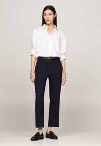 Camicia bianca con colletto, pantaloni blu tailored, cintura nera e mocassini neri. I pantaloni hanno una vestibilità cropped; la camicia presenta una tasca frontale. Texture lisce.