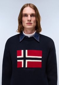 Joven con el pelo hasta los hombros que lleva un jersey azul marino con un diseño de cruz roja, blanca y negra, superpuesto a una camisa vaquera con cuello.