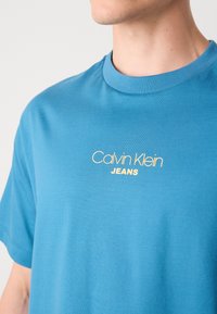 Kék pamut póló kerek nyakkal és rövid ujjal. Elöl sárga "Calvin Klein JEANS" logóval. Simább textúrával rendelkezik.