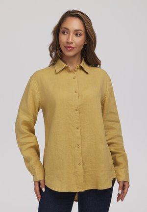 Kobieta ubrana w musztardowożółtą, długą koszulę zapinaną na guziki oraz ciemne spodnie, stojąca na jednolitym jasnoszarym tle.