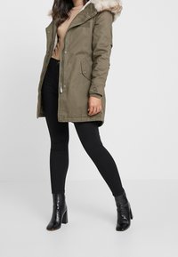 Olivgrüner Parka mit einem mit Fell gefütterten Kapuze, Frontreißverschluss und seitlichen Taschen, getragen über schwarzen Leggings und schwarzen Stiefeln mit Absatz.