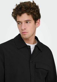 Homme aux cheveux bouclés portant une veste noire à boutons sur une chemise blanche, regardant sur le côté avec une boucle d'oreille en or dans l'oreille gauche.