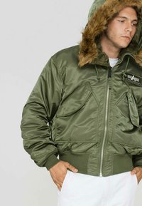 Zelen bomber jakna z zadrgo spredaj, kapuco podloženo s krznom, elastičnimi manšetami in dvema sprednjima žepoma. Material deluje sijoče in gladko.