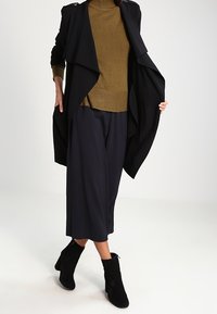 Cappotto lungo nero aperto davanti, maglione a collo alto oliva, pantaloni neri a gamba larga e stivaletti neri. Tessuto morbido, design drappeggiato e look a strati.