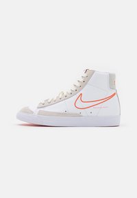 Sneaker Nike alta bianca con accenti in suede beige, logo swoosh arancione e lacci bianchi su uno sfondo bianco semplice.