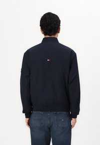 Giacca bomber blu navy con colletto rialzato, polsini e orlo elasticizzati. Presenta un piccolo dettaglio del logo rosso, bianco e blu sul retro.