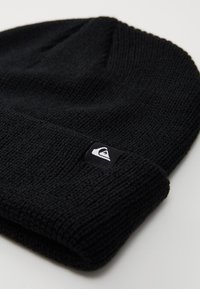 Black knitted beanie with a ribbed texture and a folded cuff. Features a small white logo tag on the cuff.