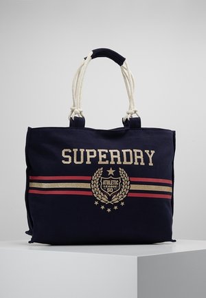 Bolso tote de lona azul marino con asas de cuerda blanca, rayas rojas y doradas, y texto y emblema "SUPERDRY Athletic League SD" en el frente.