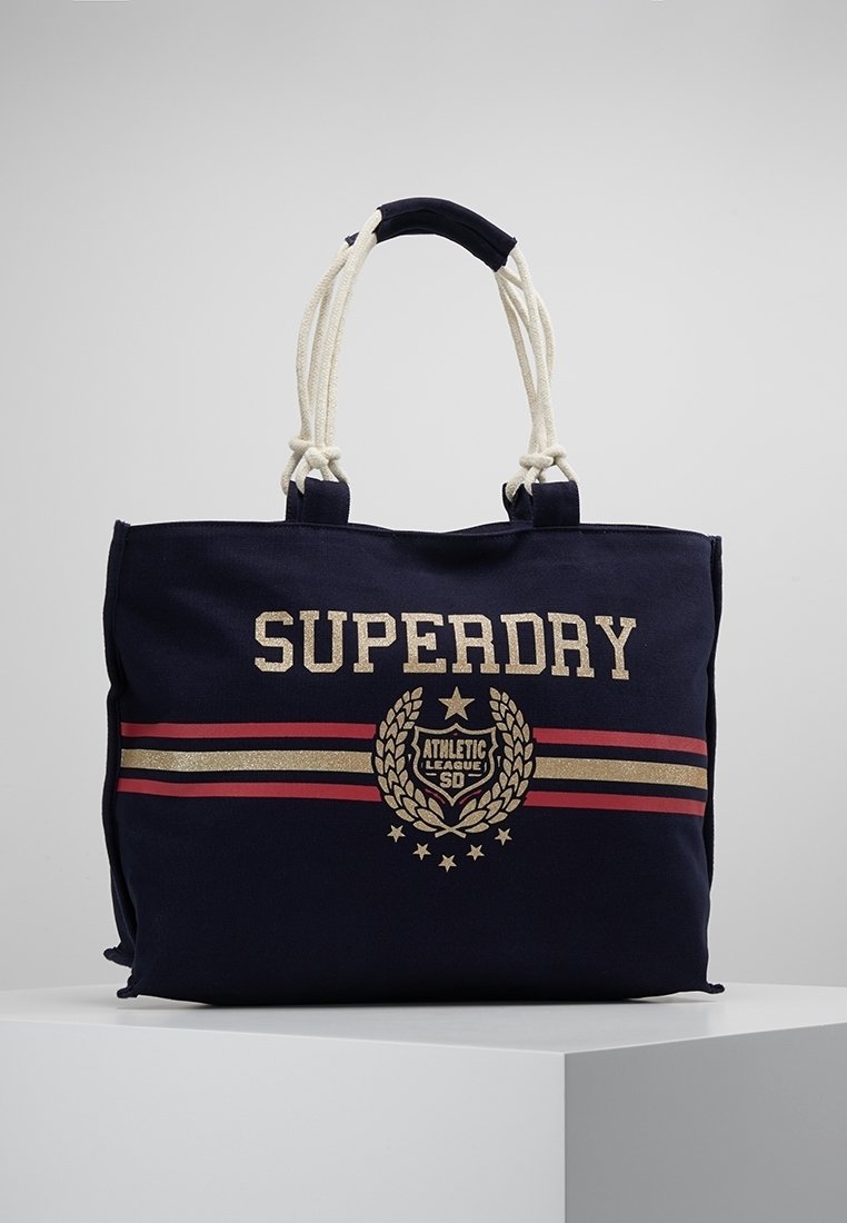 Sac fourre-tout en toile bleu marine avec des poignées en corde blanche, des rayures rouges et dorées, et le texte "SUPERDRY Athletic League SD" ainsi qu'un emblème sur le devant.