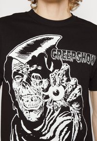 Camiseta de algodón negra con un estampado gráfico blanco que presenta un personaje de terror sosteniendo un ojo, con el texto "CREEPSHOW" en una fuente estilizada.