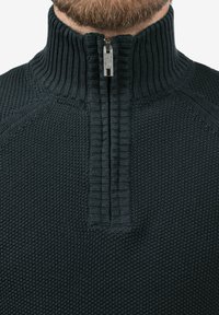 Suéter de punto oscuro con un alto cuello acanalado y un cierre de medio zip, que presenta un patrón texturizado y detalles bordados.