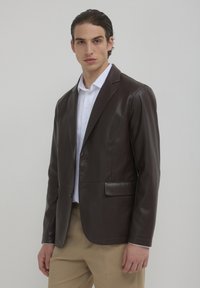 Blazer in pelle marrone con vestibilità sagomata, chiusura a un bottone e revers a punte. Abbinato a una camicia bianca e pantaloni beige.