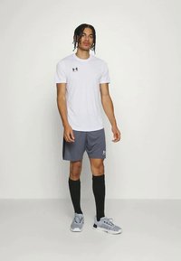 Sportiv de sex masculin îmbrăcat cu un tricou sport alb, pantaloni scurți gri, șosete negre până la genunchi și pantofi sport gri, pe un fundal simplu.