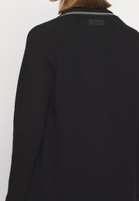 Svart stickad tröja med rundad krage och vita ränder, textil med struktur och en liten svartetikett nära halsringningen.