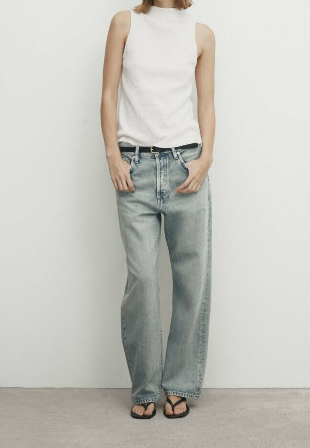 Personne portant un haut blanc sans manches, un jean bleu clair ample avec une ceinture noire, et des tongs noires, debout contre un mur blanc uni.