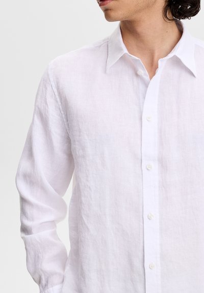 Homme aux cheveux foncés portant une chemise blanche en lin à manches longues boutonnée, vu de profil à gauche sur un fond clair uni.