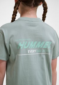 Jasnozielona bawełniana koszulka z okrągłym dekoltem. Z tyłu znajduje się napis "HUMMEL" w dynamicznej czcionce, pod nim "EVERYDAY ACTIVE" w kolorze białym.