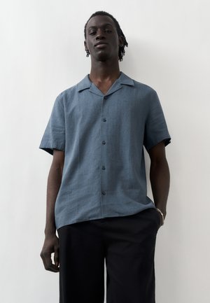 Νεαρό άτομο που φοράει μπλε κοντομάνικο πουκάμισο με κουμπιά και μαύρο παντελόνι, στέκεται μπροστά σε απλό λευκό φόντο.