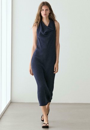 Frau läuft drinnen und trägt ein ärmelloses marineblaues Strick-Midikleid mit drapiertem Wasserfallausschnitt und schwarzen Sandalen mit Absatz.