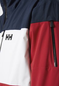 Coupe-vent bleu marine, blanc et rouge en tissu léger. Présente une poche zippée sur la manche et un grand logo "HH" sur la poitrine.