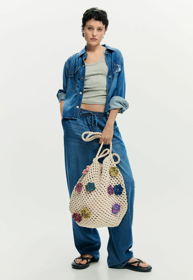 Jeune personne portant une veste en jean, un jean à cordon, un débardeur gris, tenant un grand sac en crochet crème décoré de fleurs colorées.