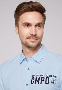 Világoskék, galléros ing, melyen hímzett "CAMP DAVID BLUE CMPD" logó található. Gombos záródás és texturált anyag jellemzi.