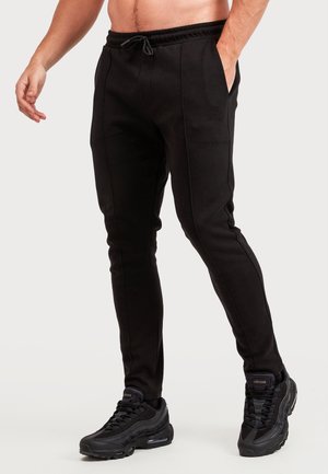 Hombre con pantalones jogger negros con bolsillos delanteros y zapatillas negras Nike Air Max, de pie con una mano en el bolsillo frente a un fondo liso.