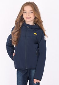 Jūrinės tamsiai mėlynos spalvos minkšta striukė su gaubtu, puikuojanti geltonu skėčio logotipu krūtinėje ir šoninėmis kišenėmis, apsivilkta su džinsais.