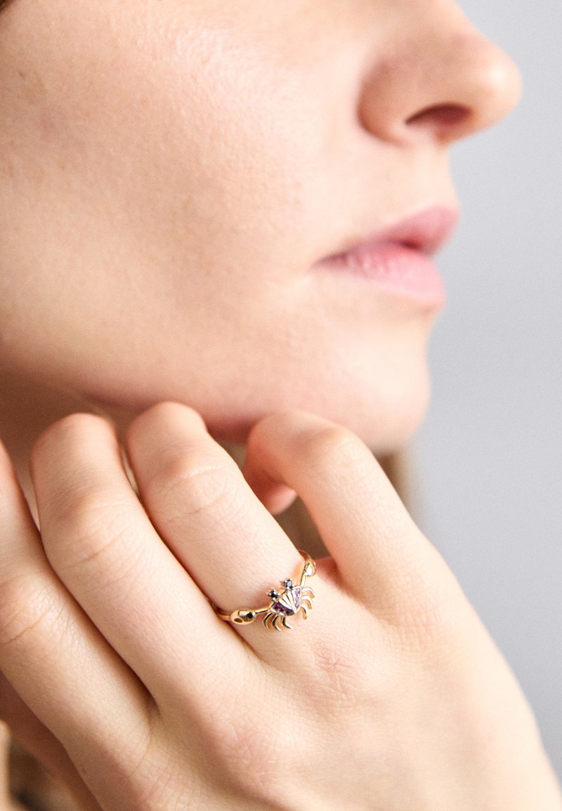 Χρυσό δαχτυλίδι σε σχήμα καβουριού με λεπτομερές σχέδιο, μπλε λεπτομέρειες και λείο περιφερειακό, που φοριέται σε χέρι που αναπαύεται στο πρόσωπο.