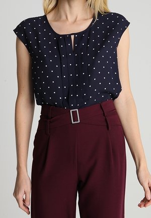 Vrouw draagt een marineblauwe mouwloze blouse met polka dots, ingestopt in een hooggetailleerde bordeauxrode broek met een riem en zilveren gesp.