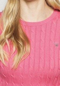 Maglione rosa a maglia con motivo a treccia, scollatura rotonda e un piccolo patch con logo sul petto. Texture morbida e design aderente.