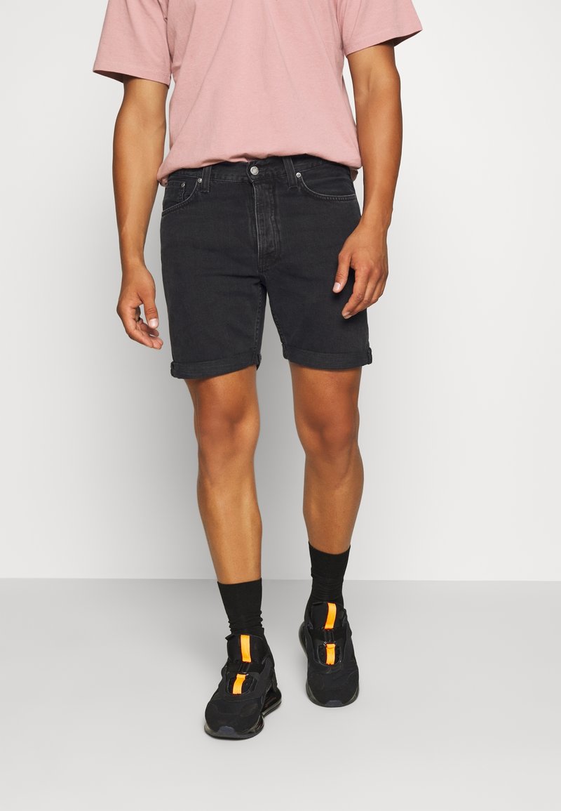 Svarta denimshorts med uppvikta kanter, tillsammans med en ljusrosa t-shirt. Modellen bär svarta sneakers med orangea detaljer och svarta strumpor.