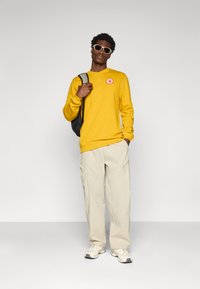 Sweatshirt jaune avec un logo circulaire, pantalon beige clair avec poches et coutures, associé à des baskets blanches et grises. Sac à dos noir et lunettes de soleil.