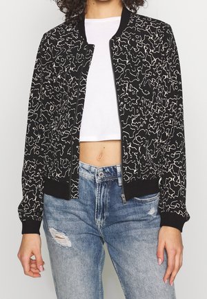 Zwarte bomberjack met een wit abstract lijnpatroon, geribde manchetten en zoom, ritssluiting, gedragen over een witte crop top en versleten denim jeans.