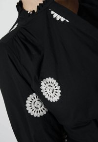 Black fabric blouse featuring embroidered white floral designs on the sleeves and a ruffled neckline detail. Soft texture and flowing design.