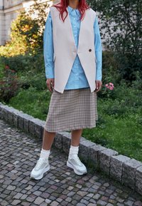 Gilet beige con un bottone sopra una camicia a righe azzurro chiaro, abbinato a una gonna a quadri marrone e sneakers bianche con dettagli menta.